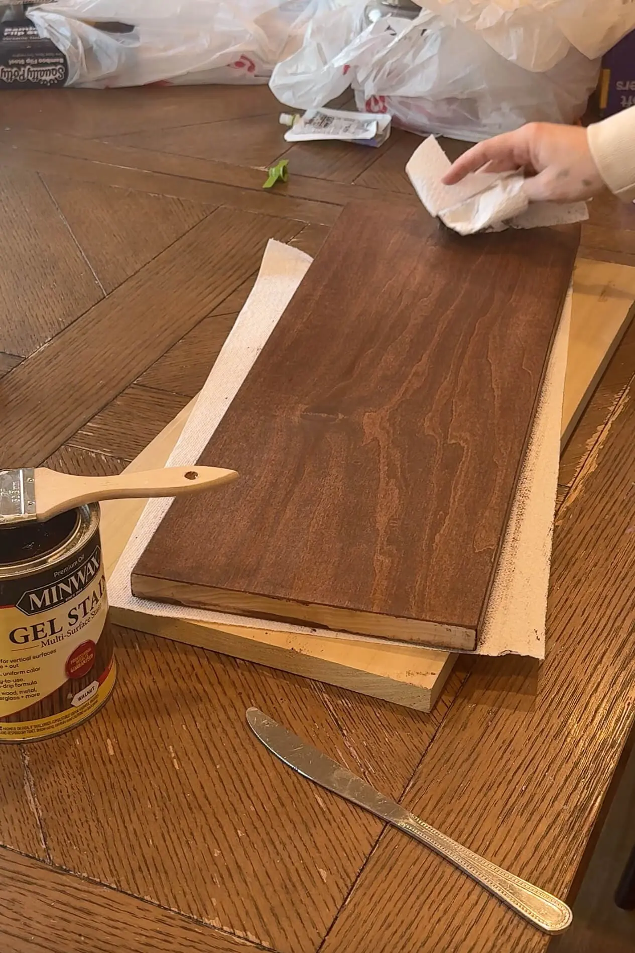 A freshly stained wooden board being wiped with a paper towel to smooth out the stain, with a can of gel stain and a brush placed on the table.